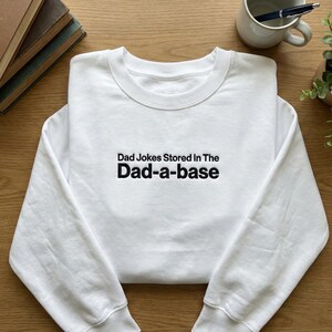 May include: A white crew neck sweatshirt with the black text "Dad Jokes Stored In The Dad-a-base". The sweatshirt has a ribbed collar, cuffs, and hem. The image is taken from a top-down perspective. The sweatshirt is made of a soft material.