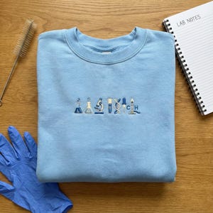 Peut inclure: Un sweat-shirt bleu clair avec des icônes brodées sur le thème de la science en nuances de bleu. Le motif comprend des béchers, un microscope, une hélice d'ADN et un brûleur de Bunsen allumé. Un bloc-notes avec "LAB NOTES" est visible.
