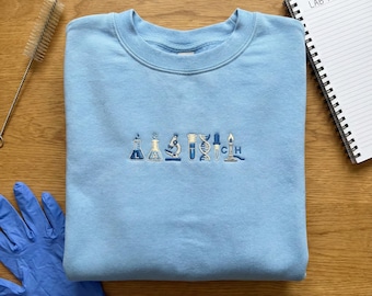 May include: A light blue sweatshirt with embroidered science-themed icons in shades of blue. The design includes beakers, a microscope, a DNA strand, and a lit Bunsen burner. A spiral notebook with "LAB NOTES" is visible.