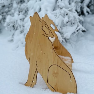 May include: Wooden cutout of a howling wolf in a snowy environment. The wolf is depicted in profile, with its head raised and mouth open. The cutout is made of light-colored wood and is partially submerged in the snow.