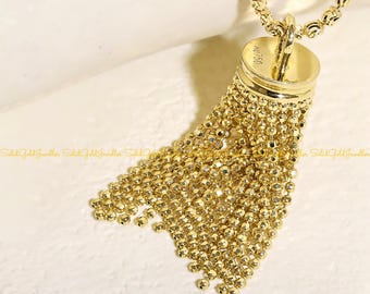 May include: A gold-colored necklace with a tassel pendant. The pendant features a cylindrical top with the text "AU750" and a cascade of small, round beads. The necklace chain is also made of small, round beads.