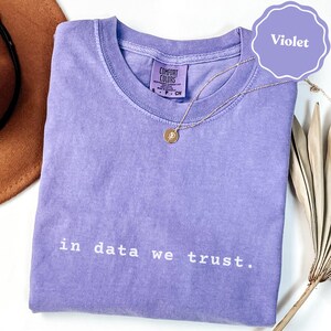 Puede incluir: Una camiseta violeta de manga corta con cuello redondo y etiqueta Comfort Colors. La prenda lleva impreso el texto blanco "in data we trust." en la parte delantera. Un fino collar dorado con un pequeño colgante circular descansa sobre la tela.