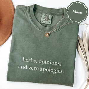 May include: A moss green crew neck t-shirt featuring the white text "herbs, opinions, and zero apologies." printed on the front. The shirt has a Comfort Colors brand label inside the collar and is paired with a delicate gold-toned chain necklace.