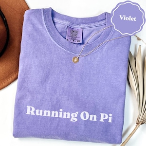 Puede incluir: Una camiseta violeta de manga corta con cuello redondo y acabado lavado vintage. El texto "Running On Pi" está impreso en blanco en el pecho. Una etiqueta de la marca Comfort Colors es visible en el cuello interior, indicando la talla S.
