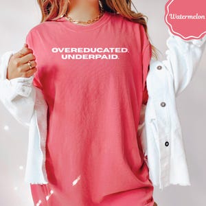 May include: A coral-colored t-shirt with the text "OVEREDUCATED. UNDERPAID." in white block letters. The shirt is worn with a white corduroy jacket. A gold chain necklace is visible.
