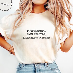 May include: An ivory short-sleeved t-shirt featuring black text centered on the chest that reads: PROFESSIONAL OVERREACTOR, LICENSED & INSURED. The shirt has a crew neckline and is paired with blue denim jeans, creating a casual and simple look.
