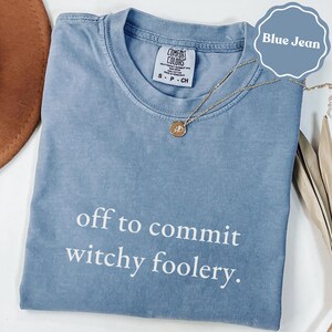 Könnte beinhalten: Ein blaues T-Shirt in Jeansoptik mit Rundhalsausschnitt und dem weißen Text "off to commit witchy foolery." auf der Vorderseite. Das Shirt hat ein Comfort Colors Markenetikett am Innenhals und wird mit einer goldenen Halskette kombiniert.