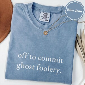 May include: A blue jean colored t-shirt with a crew neckline and a Comfort Colors brand label. The shirt features the white text "off to commit ghost foolery." A thin gold chain necklace with a small circular pendant rests on the fabric.