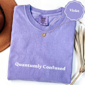 May include: A violet short-sleeved t-shirt with a crew neckline and a washed-out texture. The text "Quantumly Confused" is printed in white across the chest. A small Comfort Colors brand label is visible at the inner neck, and a gold necklace rests on top.