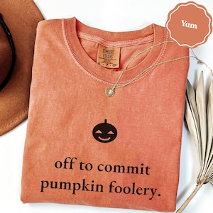 May include: A terracotta-colored short-sleeve t-shirt featuring a black graphic of a jack-o'-lantern face and the text "off to commit pumpkin foolery." The shirt has a ribbed crew neckline and a Comfort Colors brand label on the inner collar.