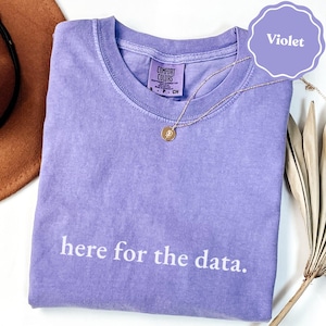 Puede incluir: Una camiseta violeta de manga corta con cuello redondo y acabado teñido en prenda. El texto 'here for the data.' está impreso en blanco en la parte delantera. Una pequeña etiqueta de la marca Comfort Colors es visible en el cuello con la talla S.