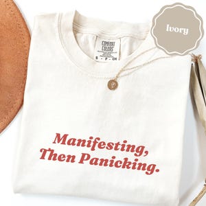 Könnte beinhalten: Ein elfenbeinfarbenes T-Shirt mit dem Text "Manifesting, Then Panicking." in dunkelroter Schrift. Das Shirt hat ein Comfort Colors Markenetikett am Halsausschnitt und wird mit einer goldfarbenen Halskette mit kleinem runden Anhänger kombiniert.