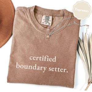 Könnte beinhalten: Ein kurzärmeliges T-Shirt in Espresso-Braun mit Rundhalsausschnitt und dem in Weiß aufgedruckten Text "certified boundary setter." auf der Vorderseite. Das T-Shirt hat ein Comfort Colors Markenetikett am Innenhals und eine silberfarbene Anhängerkette.