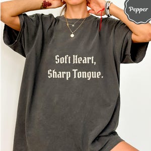 May include: A charcoal gray short-sleeve t-shirt featuring the text "Soft Heart, Sharp Tongue." printed in a light beige, gothic-style font across the chest. The garment has a relaxed, loose fit with a crew neckline and a faded, vintage-inspired wash.