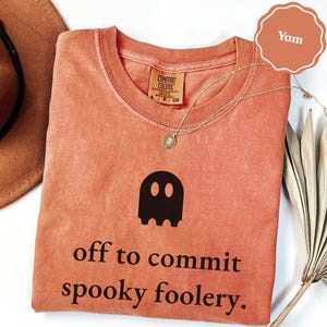 Könnte beinhalten: Ein yam-farbenes T-Shirt mit einer schwarzen Geistergrafik und dem Text "off to commit spooky foolery." Das Shirt hat einen Rundhalsausschnitt und ein Comfort Colors Markenetikett am Innenkragen. Eine dünne goldfarbene Halskette liegt auf dem Stoff.