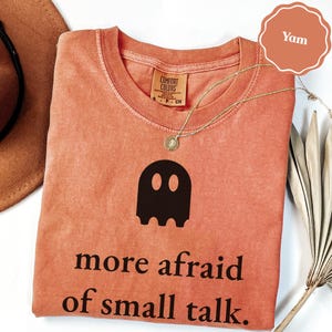 May include: A terracotta-colored t-shirt with a black graphic of a ghost and the text "more afraid of small talk." The shirt features a crew neckline, a Comfort Colors brand label, and a thin gold-toned chain necklace resting on the fabric.