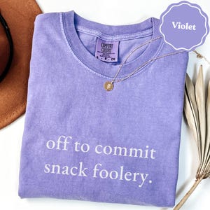 Op de afbeelding: Een violetkleurig T-shirt met korte mouwen, een ronde hals en de witte tekst "off to commit snack foolery." op de voorzijde. Het shirt is voorzien van een Comfort Colors-merklabel in de binnenkraag en heeft maat S.