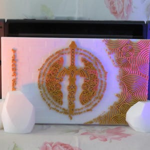 Peut inclure: Une console Nintendo Switch blanche avec une plaque frontale personnalisée représentant une épée dorée et des motifs complexes. La plaque frontale porte l'inscription "The Legend of Zelda: Tears of the Kingdom". Deux objets blancs de forme géométrique se trouvent au premier plan.