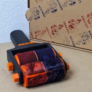 May include: A multi-color roller stamp with the word "ASTAMP" on top. The stamp has three rollers in red, purple, and orange. A cardboard box with a repeating pattern of cartoon characters and the text "424 PIZZA" is in the background.