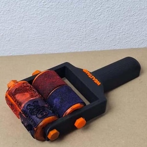 K&ouml;nnte beinhalten: Eine schwarz-orangefarbene Stempelwalze mit zwei gemusterten Walzen. Die Walzen sind rot, orange und blau. Auf dem Griff steht das Wort "WEASTAMP". Die Walze liegt auf einer beigen Oberfl&auml;che.