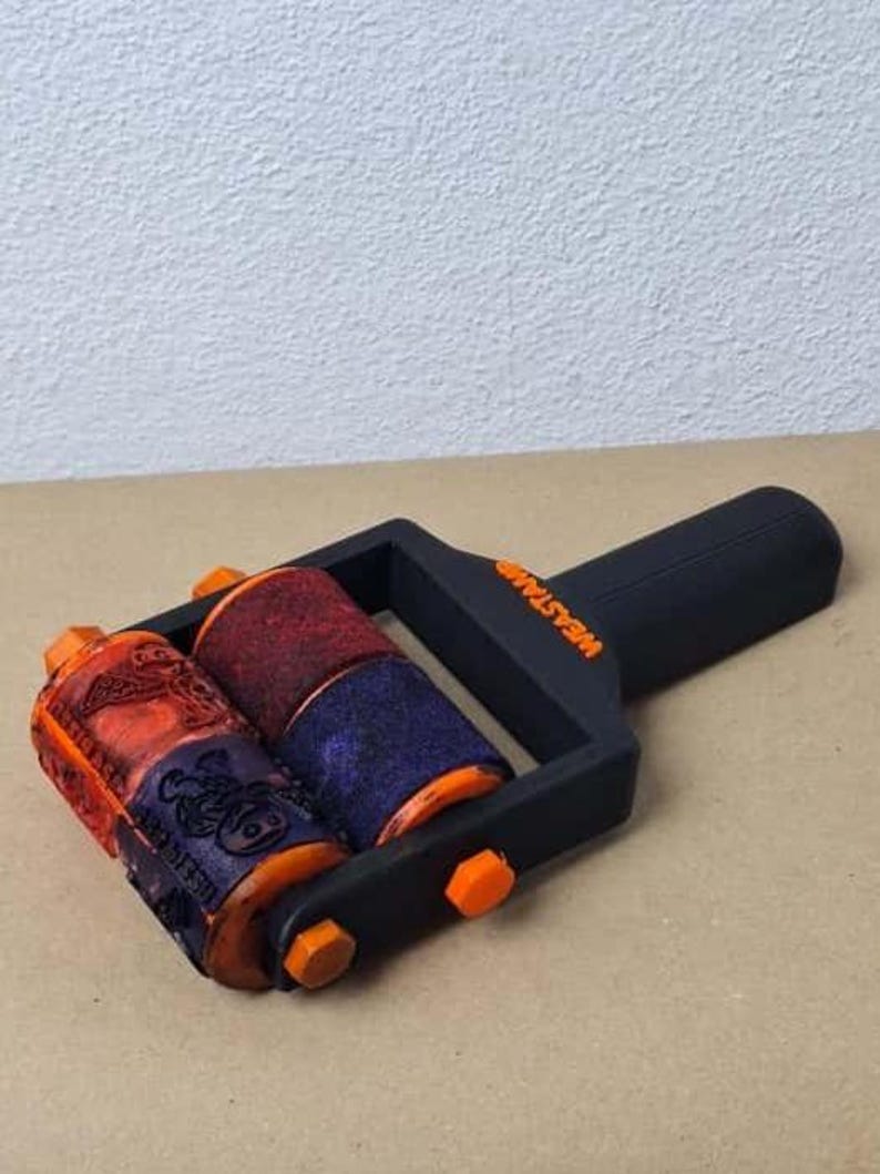 K&ouml;nnte beinhalten: Eine schwarz-orangefarbene Stempelwalze mit zwei gemusterten Walzen. Die Walzen sind rot, orange und blau. Auf dem Griff steht das Wort "WEASTAMP". Die Walze liegt auf einer beigen Oberfl&auml;che.