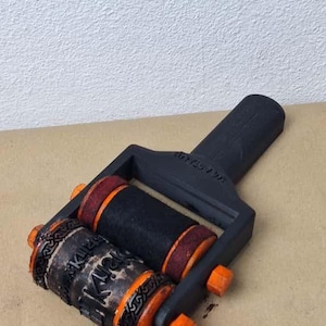 K&ouml;nnte beinhalten: Eine schwarz-orange Stempelrolle mit zwei zylindrischen Rollen. Eine Rolle hat das Wort "Kiri" in einer dekorativen Schriftart. Die andere Rolle ist schwarz mit roten Akzenten. Der Griff ist schwarz mit dem Text "2020 AW".