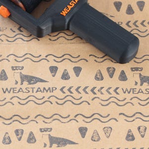 Könnte beinhalten: Ein schwarzes und orangefarbenes Weastamp-Werkzeug liegt auf braunem Papier mit einem sich wiederholenden Muster aus Ottern, Dreiecken und Wellenlinien. Das Wort "WEASTAMP" ist orange auf dem Werkzeug und schwarz auf dem Papier gedruckt.