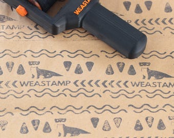 May include: A black and orange Weastamp tool rests on brown paper with a repeating pattern of otters, triangles, and wavy lines. The word "WEASTAMP" is printed in orange on the tool and in black on the paper.
