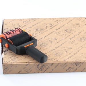 May include: A black and orange Weastamp pizza box roller stamp rests on a brown pizza box. The roller has a black roller and an orange roller. The pizza box has a repeating pattern of text and illustrations.