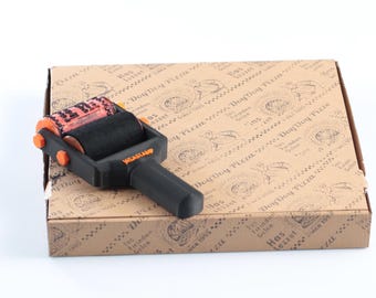 May include: A black and orange Weastamp pizza box roller stamp sits on top of a brown pizza box. The roller has a black roller and an orange roller. The pizza box has a repeating pattern of text and illustrations.