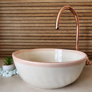 Puede incluir: Un lavabo de cerámica blanca con borde rosa, combinado con un grifo de cobre. El lavabo está sobre una superficie clara, con una pequeña planta en maceta y piedras decorativas a un lado. El grifo tiene un diseño curvo.