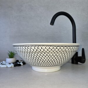 Peut inclure: Un lavabo de salle de bain en céramique blanche avec un motif géométrique noir et blanc. Le lavabo est rond et a une large bordure. Un robinet noir est visible en arrière-plan.
