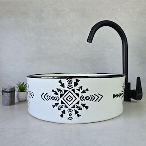 Puede incluir: Lavabo de cerámica blanca con borde negro y diseño geométrico en blanco y negro. El lavabo se muestra con un grifo negro.