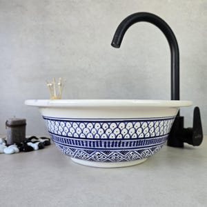 Puede incluir: Lavabo de cerámica blanca con diseño azul. El diseño presenta motivos geométricos y florales. El grifo negro es curvo. Se ven una vela y piedras decorativas.