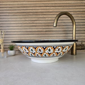 Peut inclure: Un évier en céramique blanche avec un motif floral jaune, bleu et vert. L'évier a une bordure noire et est placé sur un fond en bois.