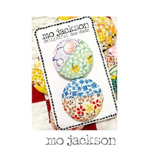 May include: Two fabric-covered buttons with floral patterns are displayed on a white card. The card has the text "mo jackson delightful doo dads". The buttons are divided into two halves with different patterns and colors. The background is a patchwork quilt.