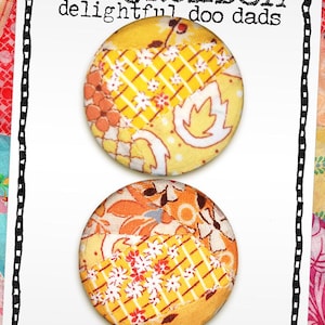 May include: Two round buttons with vintage quilt block fabric. The buttons are yellow, orange, and white, with floral and geometric patterns. The text on the card reads "mo jackson delightful doo dads" and "buttons covered with vintage quilt blocks."