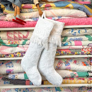 May include: Two white Christmas stockings featuring a textured, beaded surface and a decorative fringe along the top cuff. The stockings are constructed from a solid white material with a subtle, raised floral pattern throughout the fabric.