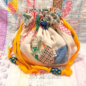 May include: A patchwork drawstring bag with a variety of fabric patterns in shades of pink, green, blue, and cream. The bag has a yellow drawstring with blue and yellow beads. The text "mo jackson" is at the bottom.