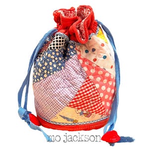 Op de afbeelding: Een patchwork trekkoordtas met een verscheidenheid aan stofpatronen, waaronder stippen, ruitjes en bloemenprints. De tas heeft een rode gefranjerde bovenkant, een blauw trekkoord en rode hartvormige knevels. De tekst "mo jackson" staat onderaan.