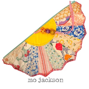 May include: A half-circle patchwork clutch with a variety of fabric patterns in shades of yellow, blue, red, and white. The clutch has a pink cord handle and decorative beaded accents. The text "mo jackson" is printed at the bottom.