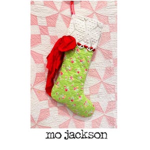 May include: A quilted Christmas stocking featuring a light green fabric with a small pink and red floral pattern. The top cuff is made of white crocheted lace, accented with a large red fabric bow and a red and white gingham hanging loop.