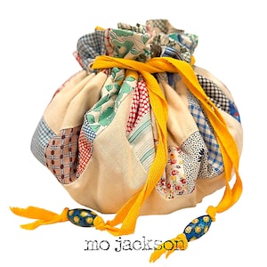 May include: A round drawstring bag with a patchwork design. The bag features a cream-colored base with colorful fabric patches in various patterns. A bright yellow ribbon cinches the top, and the text "mo jackson" is printed at the bottom.