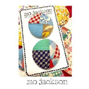 May include: Two round fabric buttons with patchwork designs in red, blue, and green, displayed on a white card. The card has the text "mo jackson dads delight" and is surrounded by a colorful floral pattern. The brand name "mo jackson" is printed at the bottom.