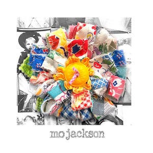 May include: A handmade fabric flower with a bright yellow center and colorful petals. The petals are made from various patterned fabrics in shades of blue, red, yellow, and pink. The text "mo jackson" is at the bottom.
