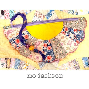 May include: A handmade, floral-patterned pouch with a yellow center and scalloped edges. The pouch features a purple zipper and handle, and a beaded tassel. The background is a patchwork quilt. The text "mo jackson" is at the bottom.