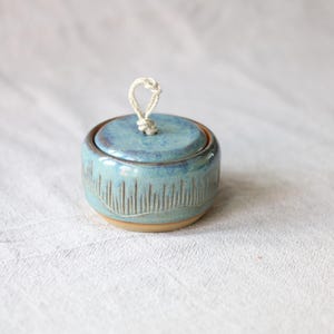 Könnte beinhalten: Ein kleines, rundes Keramikgefäß mit Deckel, das eine glänzende blaue Glasur und ein strukturiertes Muster aus vertikalen Linien an der Basis aufweist. Eine helle, geknotete Seilschlaufe ist mittig am Deckel befestigt, um diesen anzuheben.
