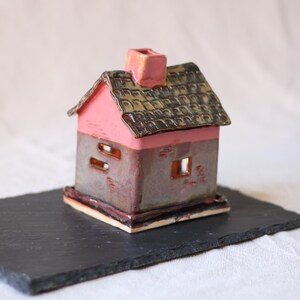 Op de afbeelding: Een klein keramieken huisje met een roze bovenkant en een donker, gespikkeld grijze onderkant. Het object heeft een getextureerd, donkerbruin pannendak met een centrale schoorsteen en kleine rechthoekige uitsparingen als ramen.