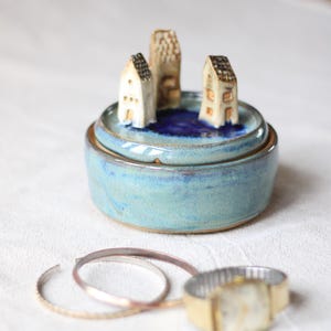 Op de afbeelding: Een ronde keramische sieradendoos met een lichtblauwe glazuurafwerking. Het deksel bevat drie kleine, met de hand gevormde witte huisjes op een donkerblauwe ronde basis. Twee dunne metalen armbanden en een goudkleurig horloge liggen ervoor.