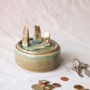 May include: A round ceramic trinket dish with a lid featuring three small, hand-sculpted house figurines on top. The dish has a glossy, muted green glaze with blue accents. The lid is decorated with a textured surface and miniature buildings.